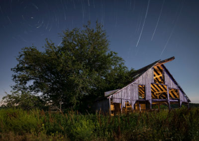 A nocturnal photograph of dilapidated barn illuminated with warm light.