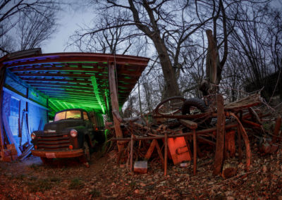 A fish-eye photograph of an abandoned farm truck and garage illuminated with colored light.
