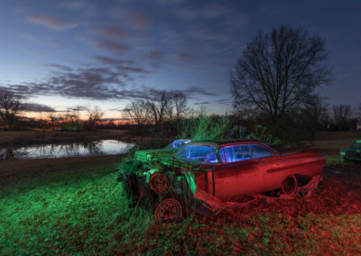 A photo during twilight of several abandoned automobiles with a pond in the background.