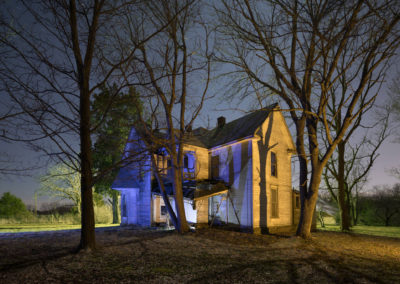 A nighttime photograph of a dilapidated home illuminated with warm and cool lights.
