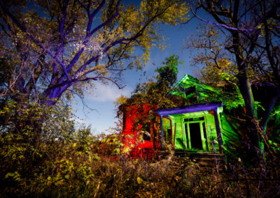 A nighttime image of an abandoned home overgrown with weeds, illuminated with red, green, and blue lights.