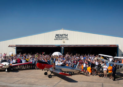 A photograph of people waving in front of a hangar with several airplanes.