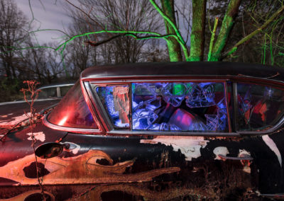 A photograph at night of an abandoned car with a cracked window illuminated with red, green, and blue lights.