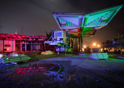 A photograph at night of a neglected gas station illuminated with red, green, and blue lights.