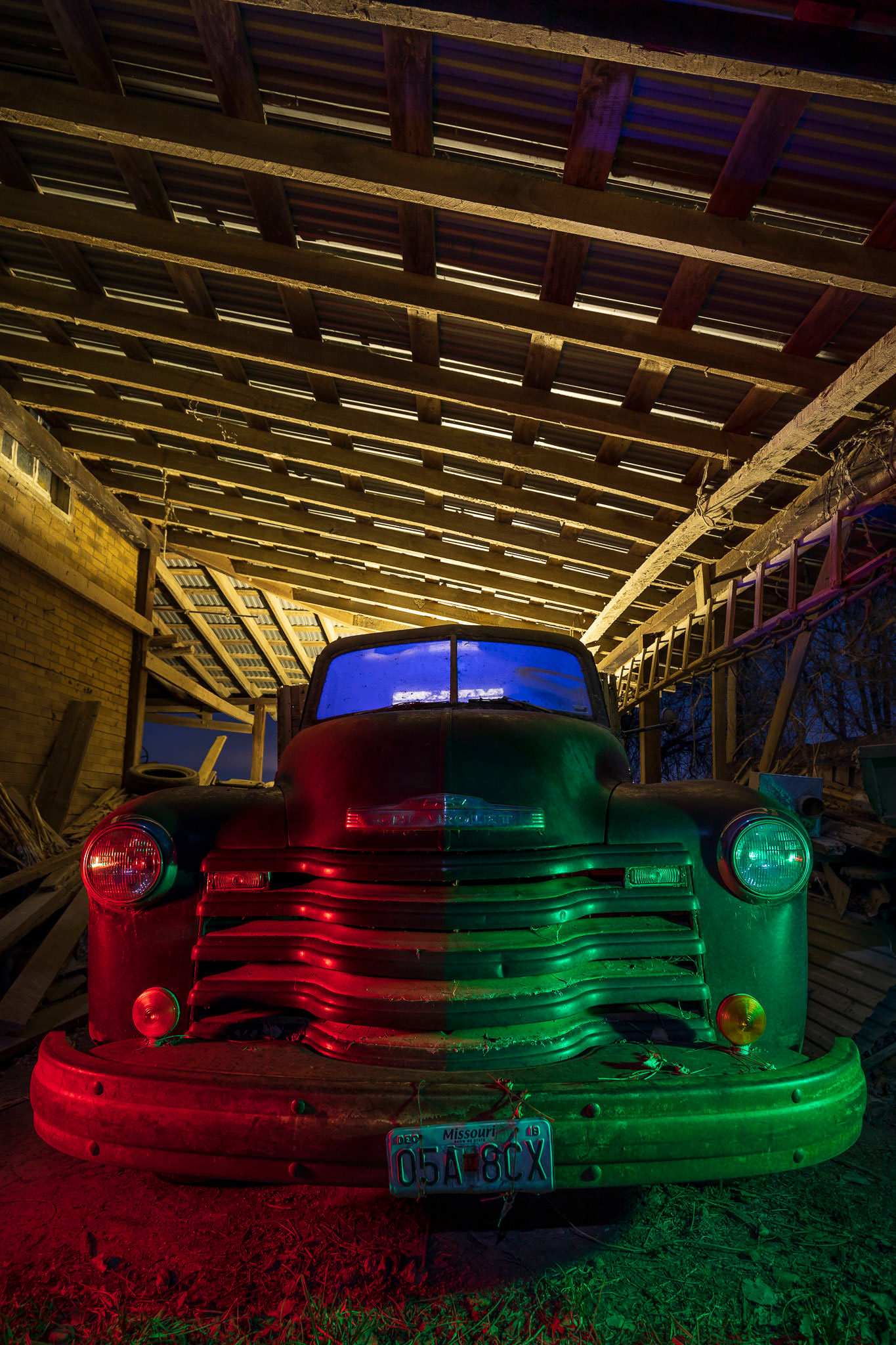 A dilapidated carport and farm truck illuminated with colored lights.