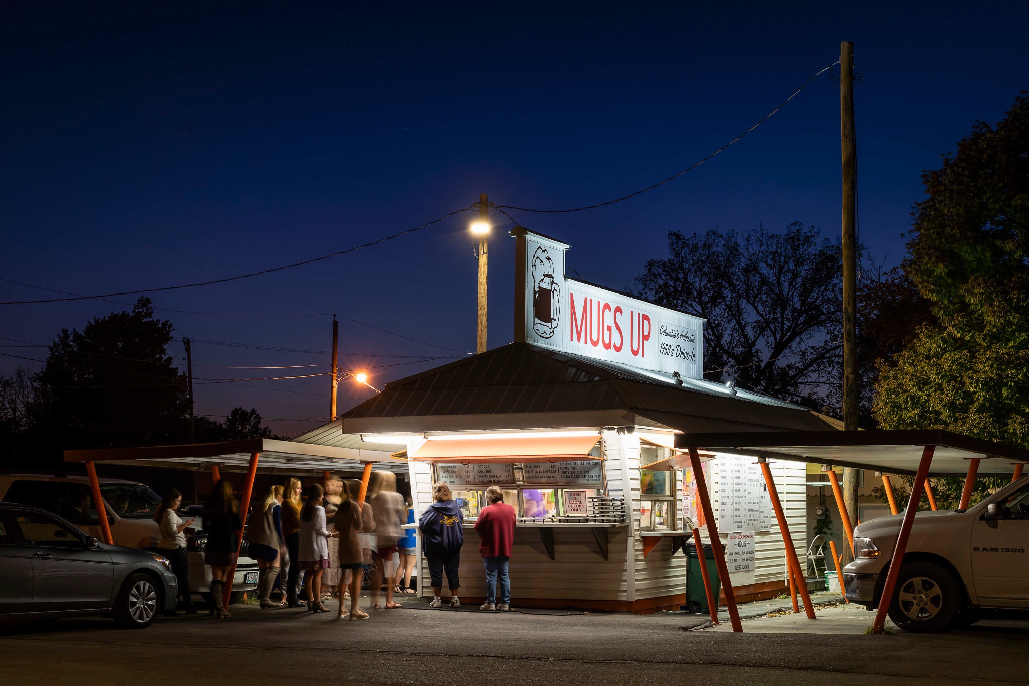 A photograph of a drive-in restaurant at twilight with customers and cars.